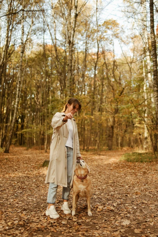 a woman standing in the woods with a dog, light brown trenchcoat, playing, profile image, snacks