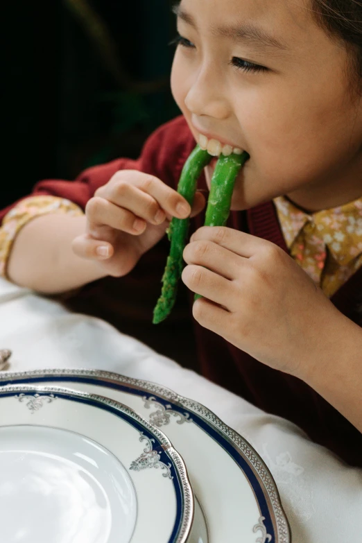 a little girl sitting at a table eating a piece of food, inspired by Elsa Beskow, unsplash, asparagus, closeup - view, made of glazed, sichuan