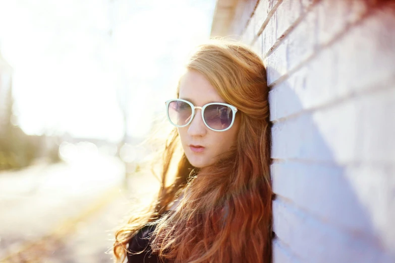 a woman leaning against a brick wall wearing sunglasses, a picture, by Julia Pishtar, unsplash, realism, long ginger hair, teenage, soft backlighting, bright sky