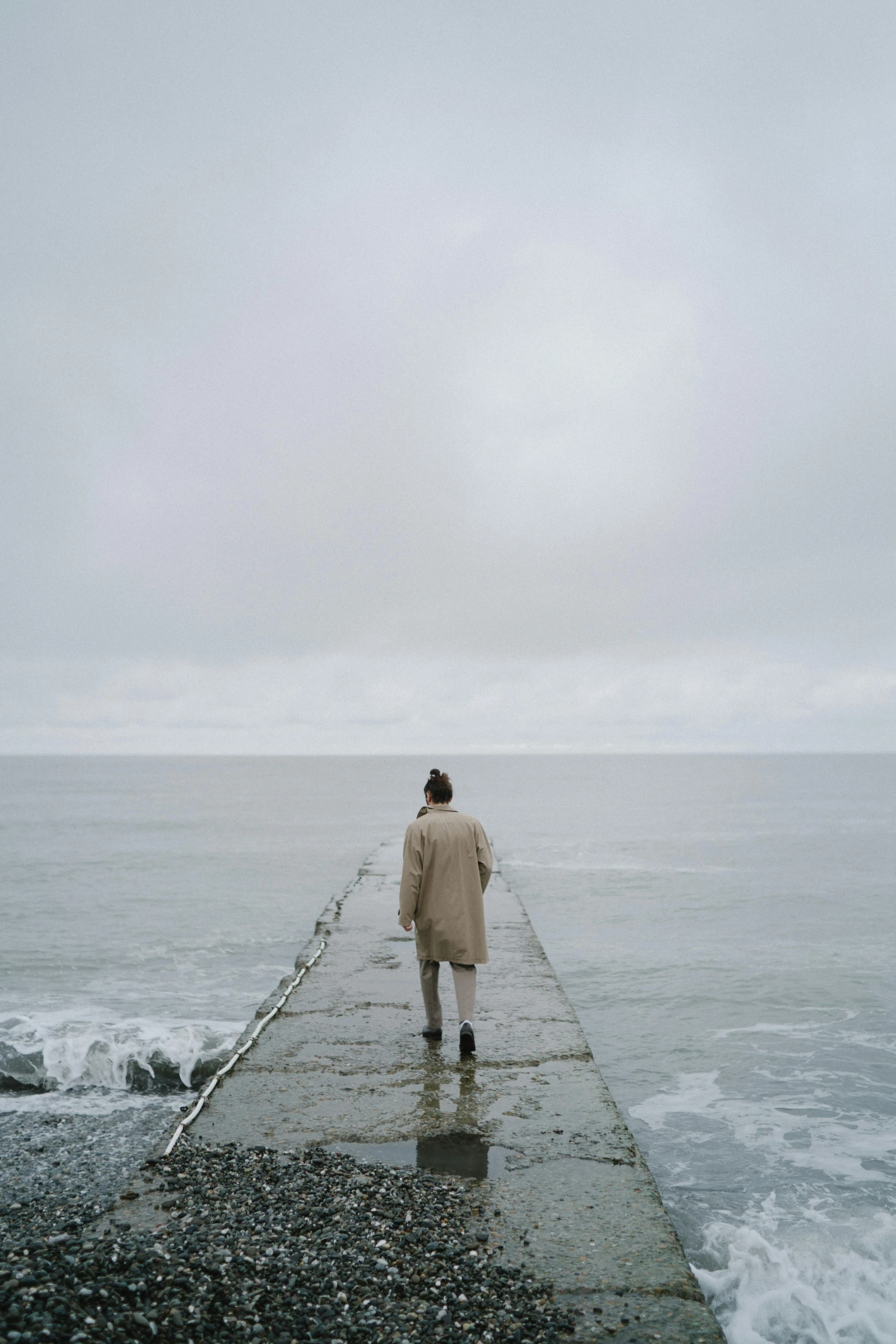 a person standing on a pier next to the ocean, gray sky, backfacing, she is wearing a wet coat, ignant