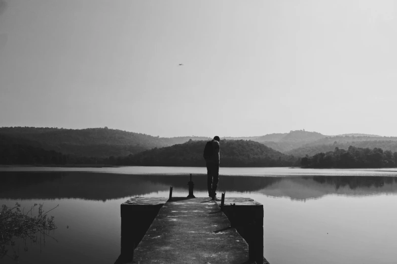 a man standing on a dock next to a lake, a black and white photo, pexels contest winner, romanticism, heartbroken, lonely astronaut, desolate :: long shot, sad men