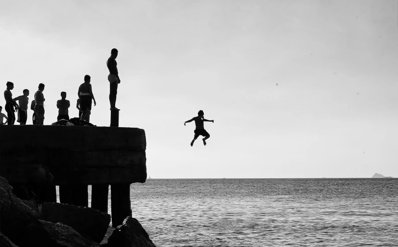 a person jumping off a cliff into the ocean, a black and white photo, by Daniel Gelon, unsplash contest winner, figuration libre, kids playing, silhouettes of people, david a trampier, eyelevel!!! view!!! photography