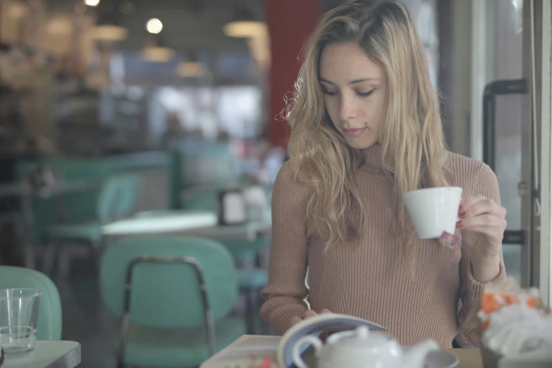 a woman sitting at a table with a cup of coffee, cinematic shot ar 9:16 -n 6 -g, reading, blonde, trending on imagestation