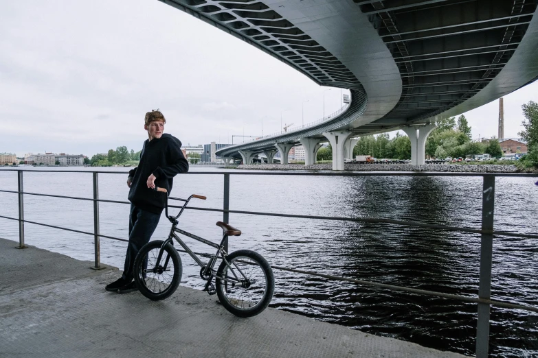 a man standing next to a bike next to a body of water, by Jaakko Mattila, sitting under bridge, he is about 2 0 years old, bulky build, action sports