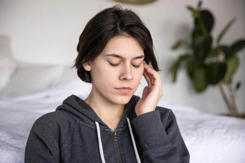 a woman sitting on a bed with her eyes closed, by Nina Hamnett, trending on pexels, hurufiyya, tinnitus, boy with neutral face, in pain, transplanted hand to head