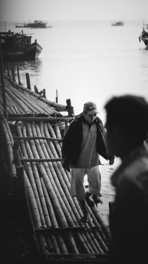 a man walking across a bamboo bridge next to a body of water, a black and white photo, an oldman, movie filmstill, candid photograph, teaching