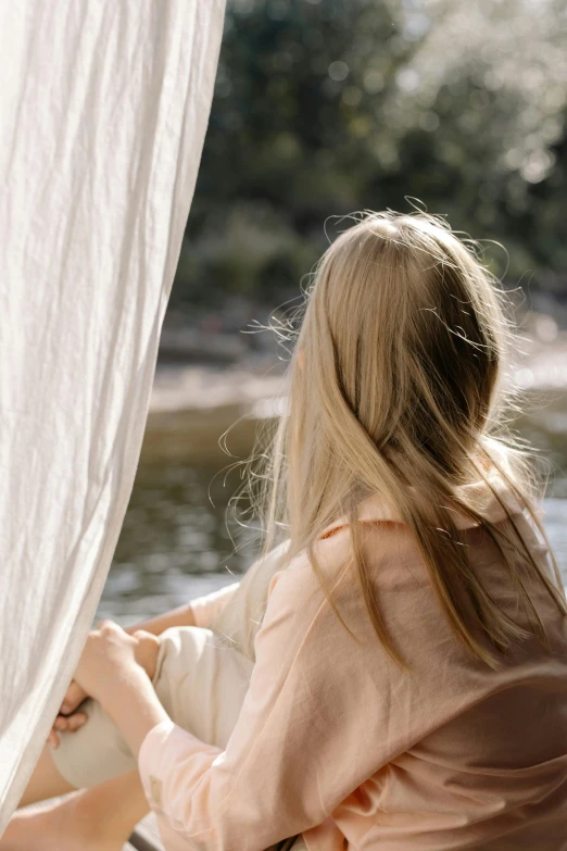 a woman sitting in a boat looking out at the water, white cloth in wind shining, a girl with blonde hair, natural muted tones, fully covered in drapes