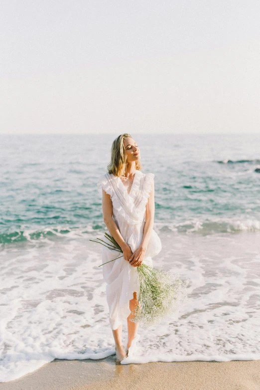 a woman standing on top of a beach next to the ocean, holding flowers, wearing white dress, pale skin curly blond hair, clean glow