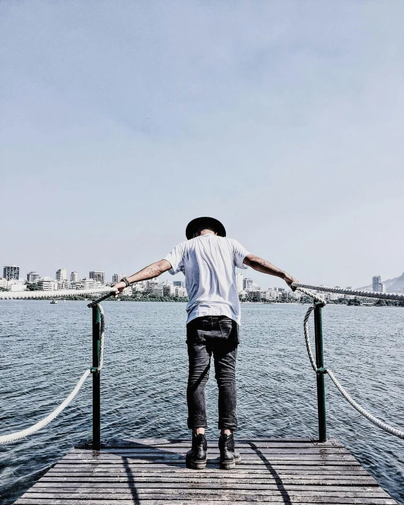 a man standing on a dock next to a body of water, pexels contest winner, happening, looking over city, pose(arms up + happy), androgynous person, about to step on you