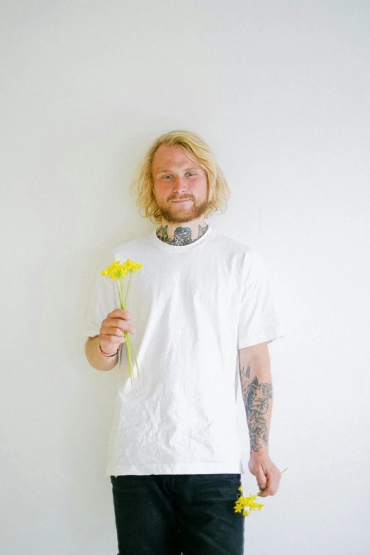 a man standing in front of a white wall holding a yellow flower, an album cover, inspired by Jesper Ejsing, unsplash, dressed in a white t-shirt, one man is blond, felix englund, slightly smiling