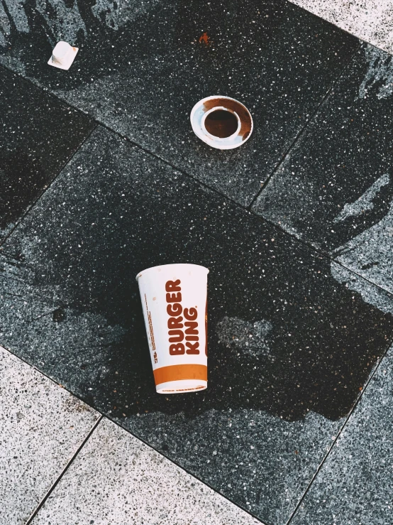 a cup of coffee sitting on top of a sidewalk, by Attila Meszlenyi, burger king, orange and white, wet floors, grunge aesthetic