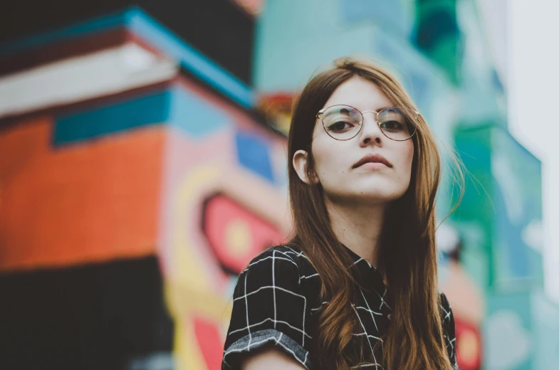 a woman with glasses standing in front of a building, trending on unsplash, graffiti, girl with brown hair, square rimmed glasses, multicolored, avatar image