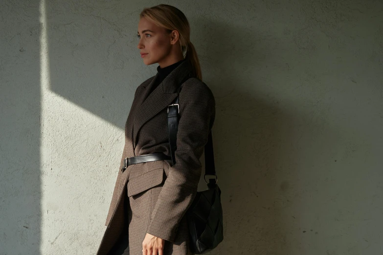 a woman standing against a wall with a handbag, by Nina Hamnett, minimalism, black and brown, wearing hunter coat, thumbnail, afternoon lighting