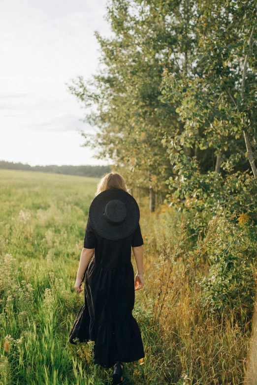 a woman walking through a field of tall grass, unsplash, wearing black old dress and hat, next to a tree, lush countryside, facing away