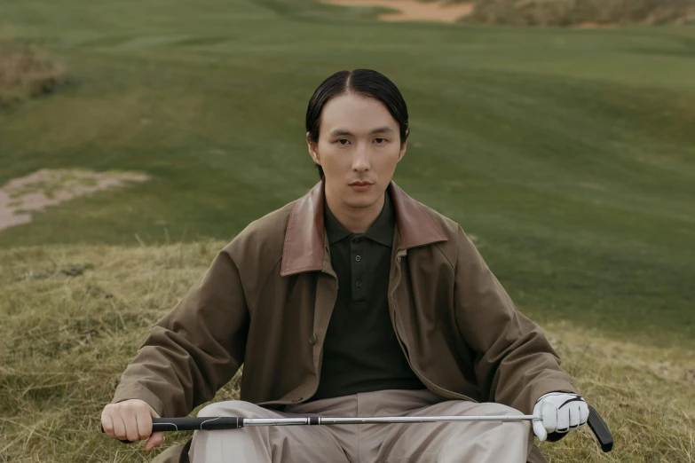 a man sitting on top of a grass covered field, an album cover, inspired by Fei Danxu, golf course, brown clothes, androgynous male, portrait featured on unsplash