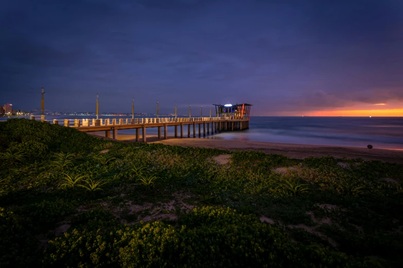 a pier sitting on top of a sandy beach next to the ocean, by Peter Churcher, unsplash contest winner, australian tonalism, blue hour, vibrant lights, south african coast, slide show