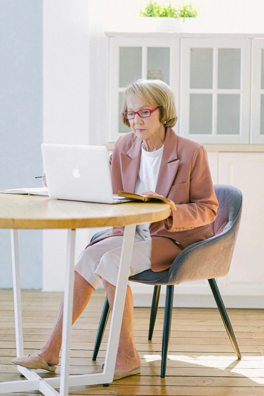 a woman sitting at a table using a laptop computer, inspired by Alexander Roslin, pexels, wearing a light - pink suit, older woman, hr ginger, sitting on designer chair