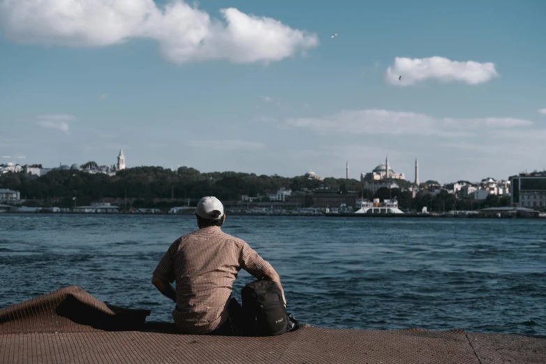 a man sitting next to a body of water, by Ismail Acar, pexels contest winner, hurufiyya, istanbul, brown, from a distance, [ cinematic