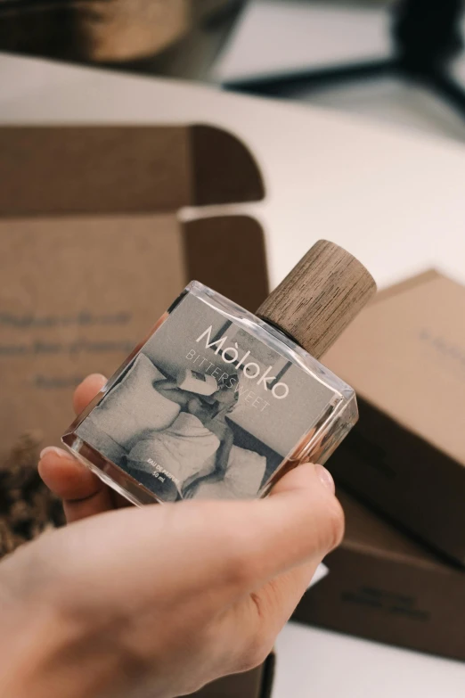 a person holding a bottle of perfume in front of a box, detailed product image, mulato, grey, cardboard