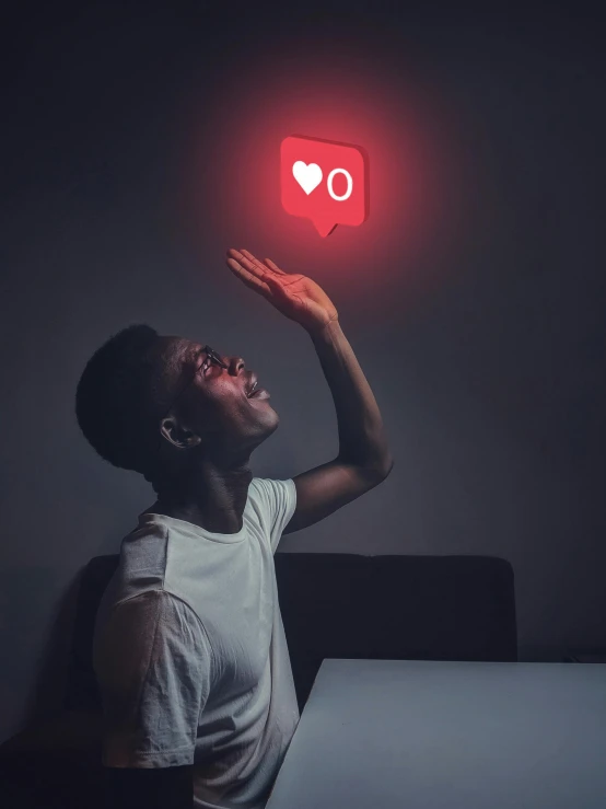 a person sitting at a table with a red light above their head, a hologram, inspired by Carrie Mae Weems, trending on pexels, heart eyes, telegram sticker, black teenage boy, rgb wall light