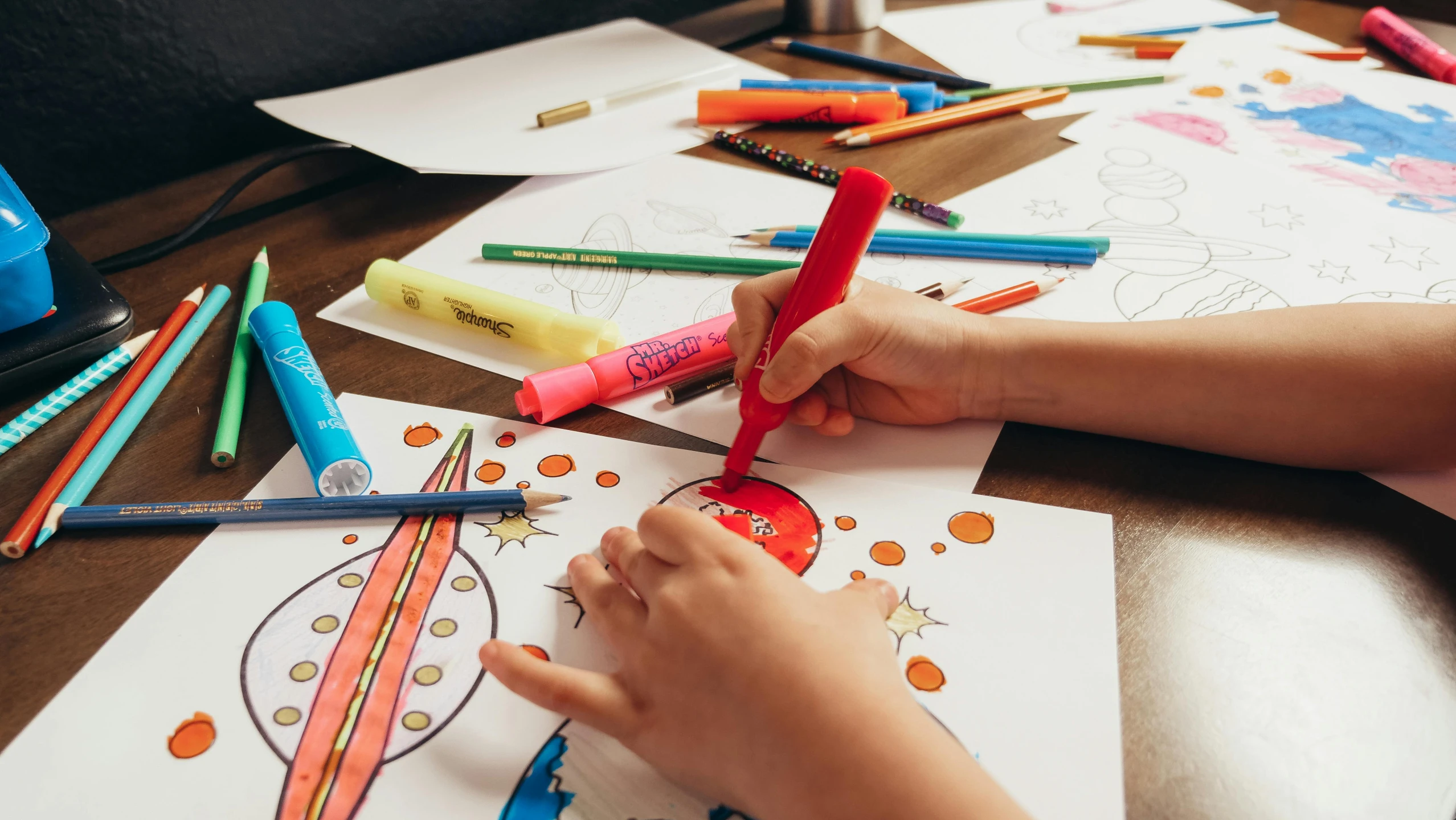 a child is drawing with colored crayons on a table, by Nicolette Macnamara, pexels contest winner, rainbow coloured rockets, marvel comic book drawing, ornate designs on desk, brand colours are red and blue