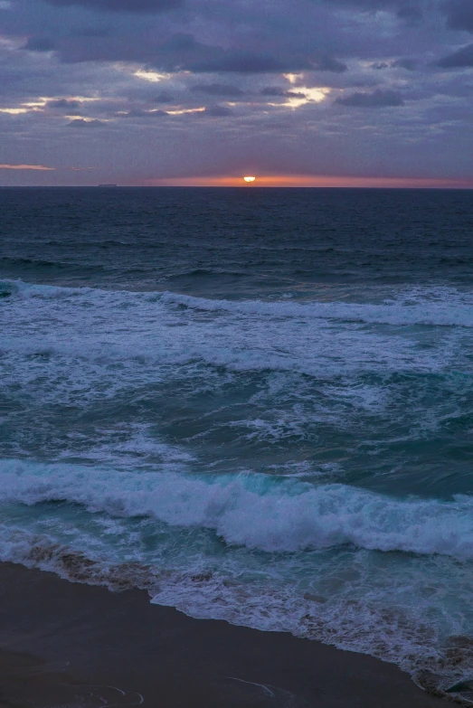 a man standing on top of a beach next to the ocean, purple sunset, in rough seas with large waves, manly, the emerald coast