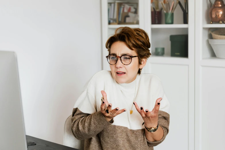 a woman sitting at a desk in front of a computer, by Emma Andijewska, pexels contest winner, he is wearing a brown sweater, arguing, wearing black frame glasses, on a white table