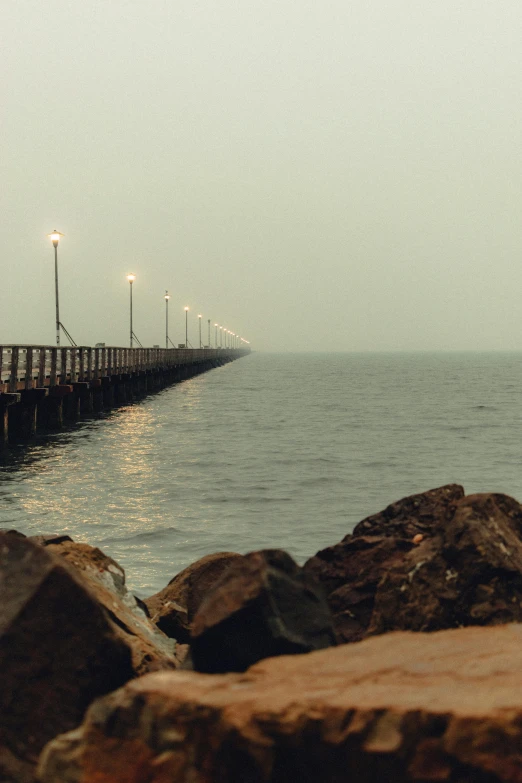 a pier in the middle of a body of water, moody hazy lighting, rocky coast, looking off into the distance, brown