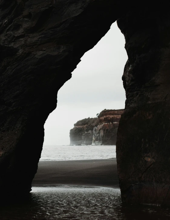 a person standing in a cave on a beach, grey skies, background image, gothic arch frame, black sand