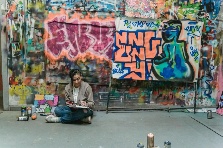 a man sitting on the ground in front of a wall covered in graffiti, in her art room, underground art, illustration »