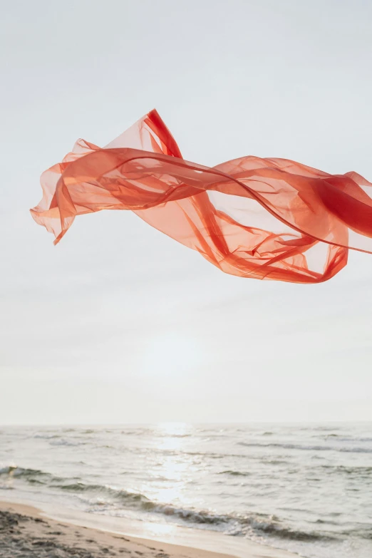 a woman standing on top of a sandy beach next to the ocean, inspired by Christo, romanticism, swirling red-colored silk fabric, light red and orange mood, flying over the ocean, ribbon