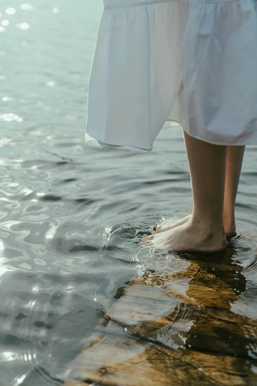 a person standing on a rock in the water, wearing a white dress, barefeet, upclose, wearing a robe