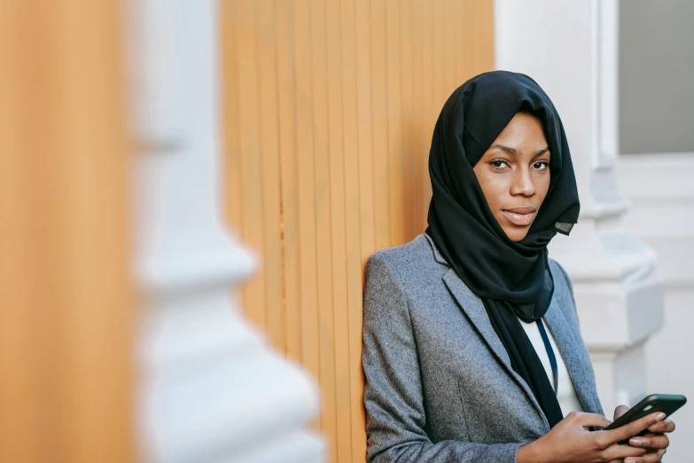 a woman in a hijab looking at her cell phone, trending on unsplash, hurufiyya, wearing a blazer, looking serious, leaning on door, black woman