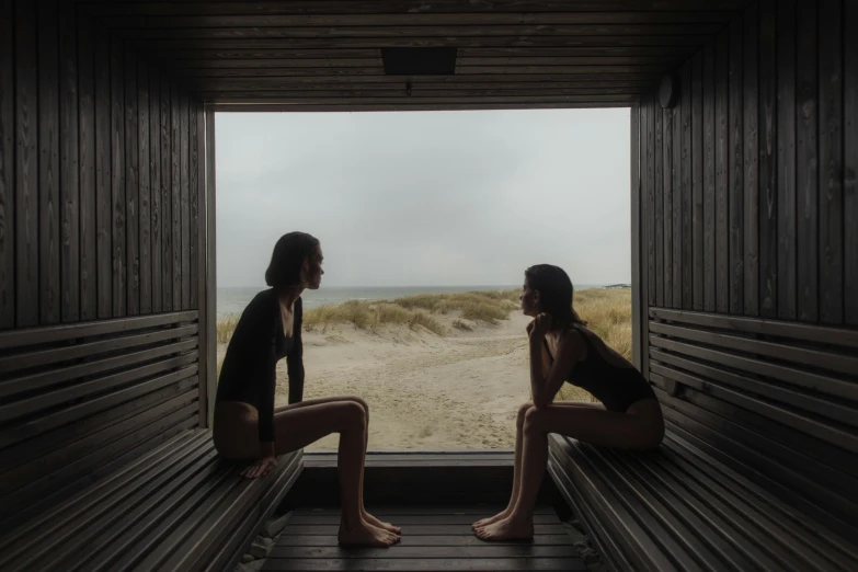 a couple of women sitting on top of a wooden bench, by Emma Andijewska, unsplash contest winner, sauna, inside in a glass box, beaches, facing each other