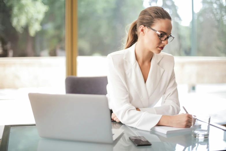 a woman sitting at a table in front of a laptop computer, by Nicolette Macnamara, pexels contest winner, royal commission, writing on a clipboard, wearing white suit and glasses, worksafe. instagram photo