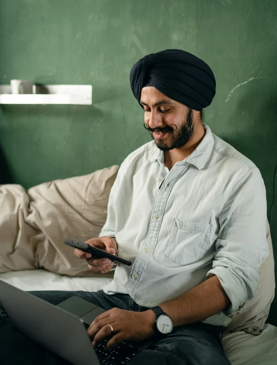 a man sitting on a bed using a laptop, inspired by Manjit Bawa, trending on pexels, renaissance, holds a smart phone in one hand, giddy smirk, turban, thumbnail