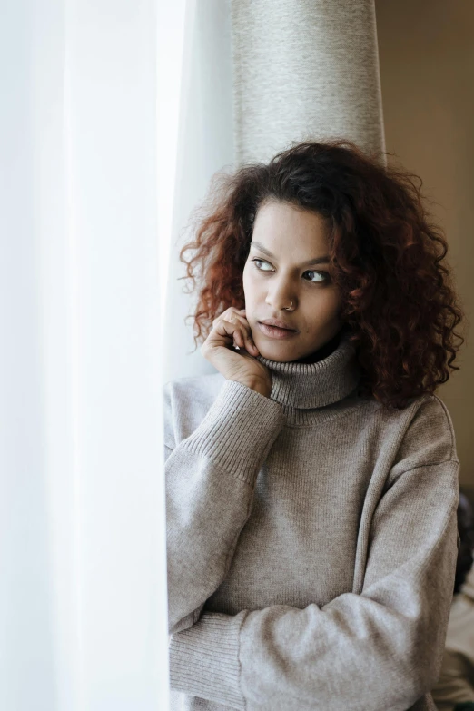 a woman standing in front of a window talking on a cell phone, a portrait, trending on unsplash, renaissance, brown sweater, looking melancholy, curly, wearing turtleneck