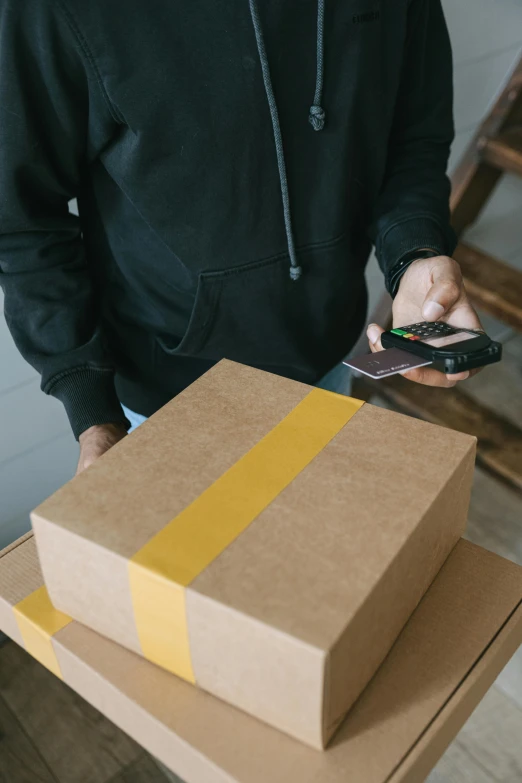 a man standing next to a stack of boxes holding a cell phone, trending on pexels, detailed product image, black and yellow tracksuit, brown, postage