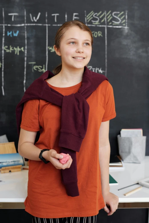 a young woman standing in front of a blackboard, a picture, shutterstock contest winner, wearing an orange t shirt, wearing a scarf, maroon, young teen