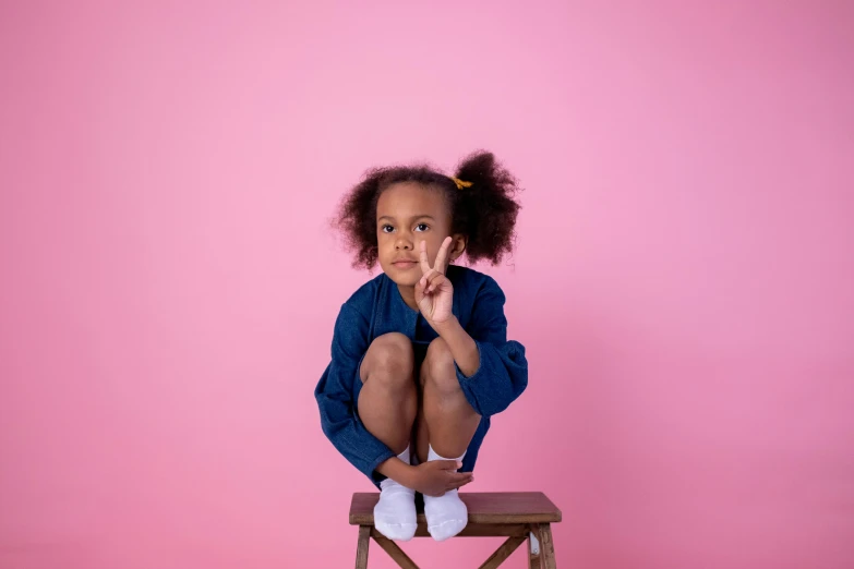 a little girl sitting on top of a wooden chair, by Sophia Beale, pexels contest winner, peace sign, black young woman, blue and pink shift, sarcastic pose