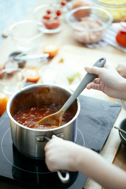 a person stirring food in a pot on a stove, tomato sauce, subtle detailing, ingredients on the table, thumbnail