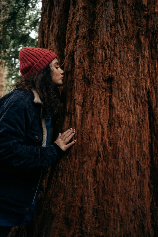 a woman leaning against a tree in a forest, by Jessie Algie, unsplash contest winner, environmental art, hat and hoodie, giant sequoia, profile image, wearing red jacket