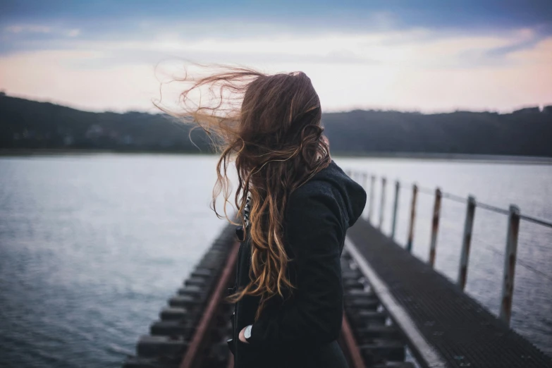 a woman standing on a train track next to a body of water, trending on pexels, long windy hair style, background blurred, what depression looks like, near a jetty