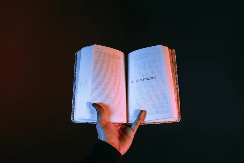 a person holding an open book in their hand, by Carey Morris, pexels, hyperrealism, with neon lighting, biblical, with a black background, instagram post