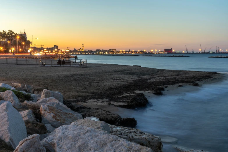 a group of rocks sitting on top of a beach next to a body of water, by Carlo Martini, pexels contest winner, graffiti, vista of a city at sunset, port, spanish, near a jetty