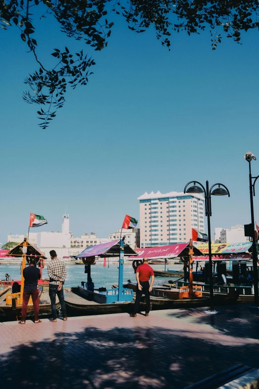 a group of people walking down a sidewalk next to a body of water, hurufiyya, colorful buildings, dubai, blue skies, 4k photo”