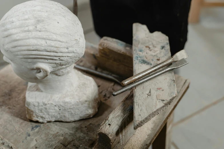 a statue sitting on top of a wooden table, a marble sculpture, inspired by Diego Giacometti, trending on unsplash, working in the forge, close - up on face, metal readymade, white clay