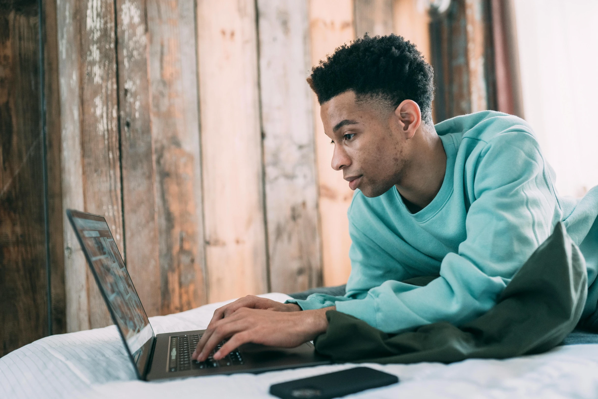 a man laying on a bed using a laptop, trending on pexels, with teal clothes, black teenage boy, looking smart, from waist up