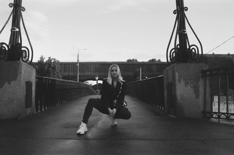 a black and white photo of a woman squatting on a bridge, by Christen Dalsgaard, wearing a tracksuit, posing for a picture, low quality footage, doing a kick