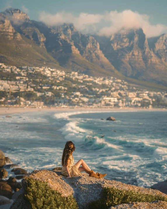 a woman sitting on top of a rock next to the ocean, pexels contest winner, south african coast, with a city in the background, conde nast traveler photo, beach aesthetic